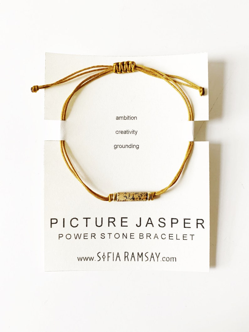 May include: A gold cord bracelet with a brown and white picture jasper stone bead. The bracelet is tied with a knot and has the words "ambition, creativity, grounding" written on the card. The card also says "Picture Jasper Power Stone Bracelet" and "www.sofiaramsay.com".