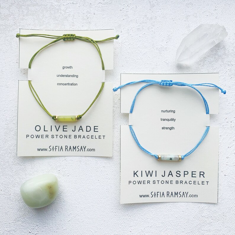 May include: Two power stone bracelets on white cards. The green bracelet has an olive jade stone and the text "growth, understanding, concentration, OLIVE JADE, POWER STONE BRACELET, www.SOFIA RAMSAY.com". The blue bracelet has a kiwi jasper stone and the text "nurturing, tranquility, strength, KIWI JASPER, POWER STONE BRACELET, www.SOFIA RAMSAY.com".