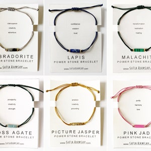 May include: Six different power stone bracelets with various coloured cords and stones. Each bracelet has a card with the stone name, properties, and website address. The stones are Labradorite, Lapis, Malachite, Moss Agate, Picture Jasper, and Pink Jade.