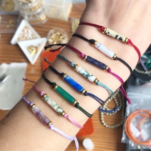 May include: A wrist with eight colourful bracelets featuring different coloured stones and beads. The bracelets are made with thin cords and have a simple design.
