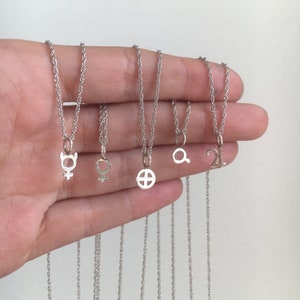 May include: Five silver necklaces with different charms, including a female symbol, a cross, a magnifying glass, and a number 21. The necklaces are on a hand, and the charms are visible.