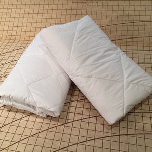 Puede incluir: Dos protectores de colchón acolchados blancos con costuras en forma de diamante. Los protectores están doblados y colocados sobre una superficie cuadriculada marrón.