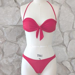 May include: A red bikini set displayed on a mannequin. The top is a bandeau style with a tie in the center, and the bottoms are a classic cut. The mannequin is set against a textured stone wall.