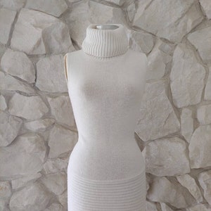 May include: A sleeveless, off-white turtleneck dress displayed on a vintage dress form. The dress features a ribbed turtleneck and a horizontally ribbed skirt section. The dress is made of a soft, textured knit fabric.