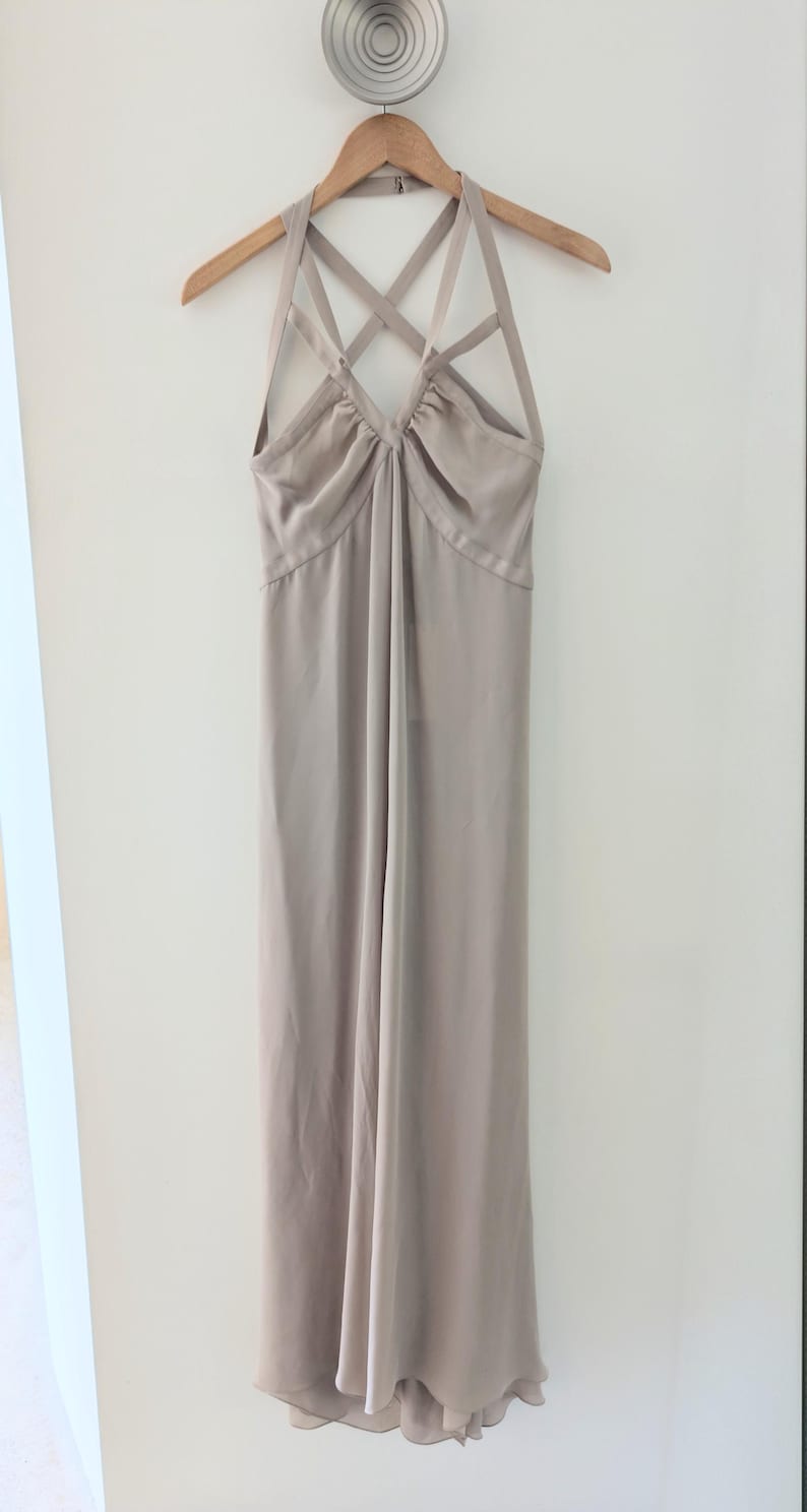 May include: A floor-length, light taupe dress featuring a halter neckline with multiple crisscrossing straps across the chest. The bodice is gathered at the center, leading into a long, flowing skirt that hangs straight down from the waistline.