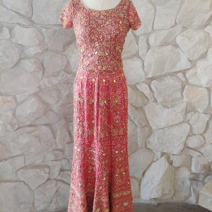 May include: A red formal gown with intricate gold embroidery and embellishments. The dress features short sleeves and a floor-length skirt, displayed on a mannequin against a stone wall background. The bodice and skirt are covered in detailed patterns.