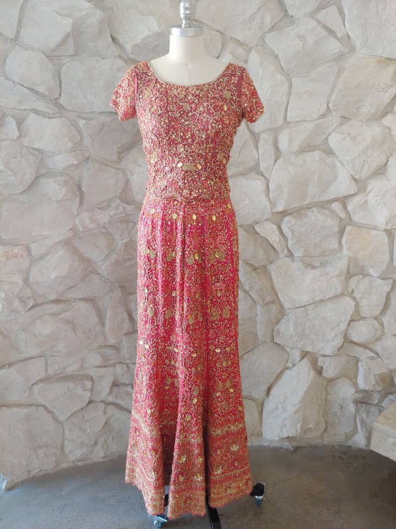 May include: A red formal gown with intricate gold embroidery and embellishments. The dress features short sleeves and a floor-length skirt, displayed on a mannequin against a stone wall background. The bodice and skirt are covered in detailed patterns.