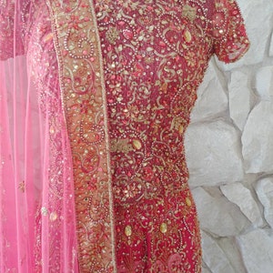 May include: A vibrant, ornate, red and gold embroidered dress with a matching pink sheer scarf. The dress features intricate beadwork and embellishments, creating a luxurious and detailed appearance. The short-sleeved dress is displayed on a mannequin.