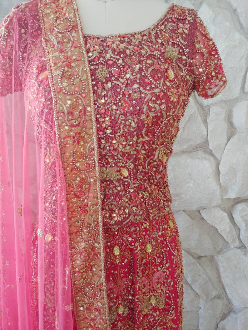 May include: A vibrant, ornate, red and gold embroidered dress with a matching pink sheer scarf. The dress features intricate beadwork and embellishments, creating a luxurious and detailed appearance. The short-sleeved dress is displayed on a mannequin.