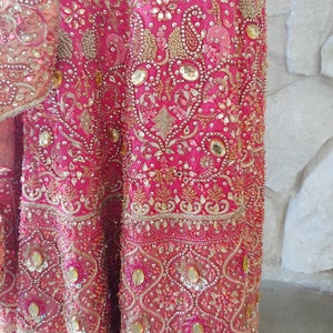 May include: Close-up of a vibrant pink garment, heavily embellished with gold embroidery, sequins, and beads. The intricate design features floral and paisley patterns. The fabric has a luxurious sheen, suggesting a formal or ceremonial attire.
