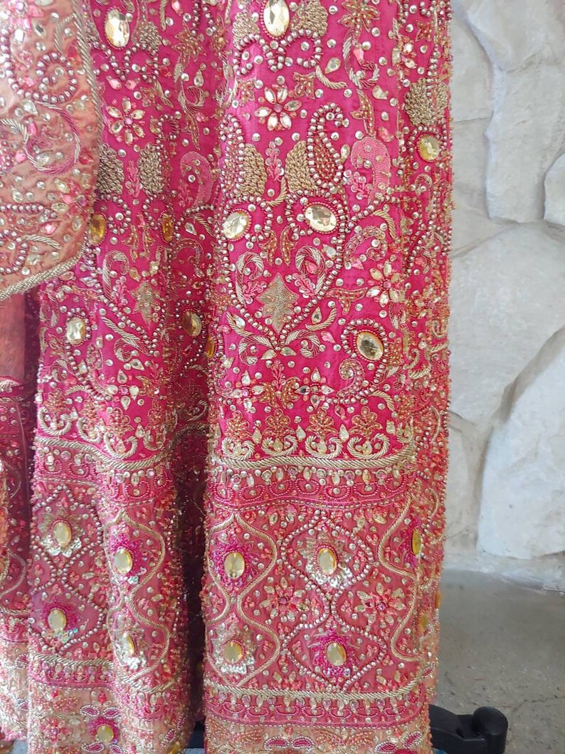 May include: Close-up of a vibrant pink garment, heavily embellished with gold embroidery, sequins, and beads. The intricate design features floral and paisley patterns. The fabric has a luxurious sheen, suggesting a formal or ceremonial attire.