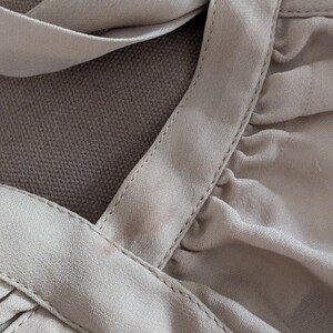 May include: A light beige garment featuring a rounded neckline with thick, stitched fabric bands that cross over each other. The fabric is gathered and pleated, creating soft folds and a textured appearance across the surface of the piece.