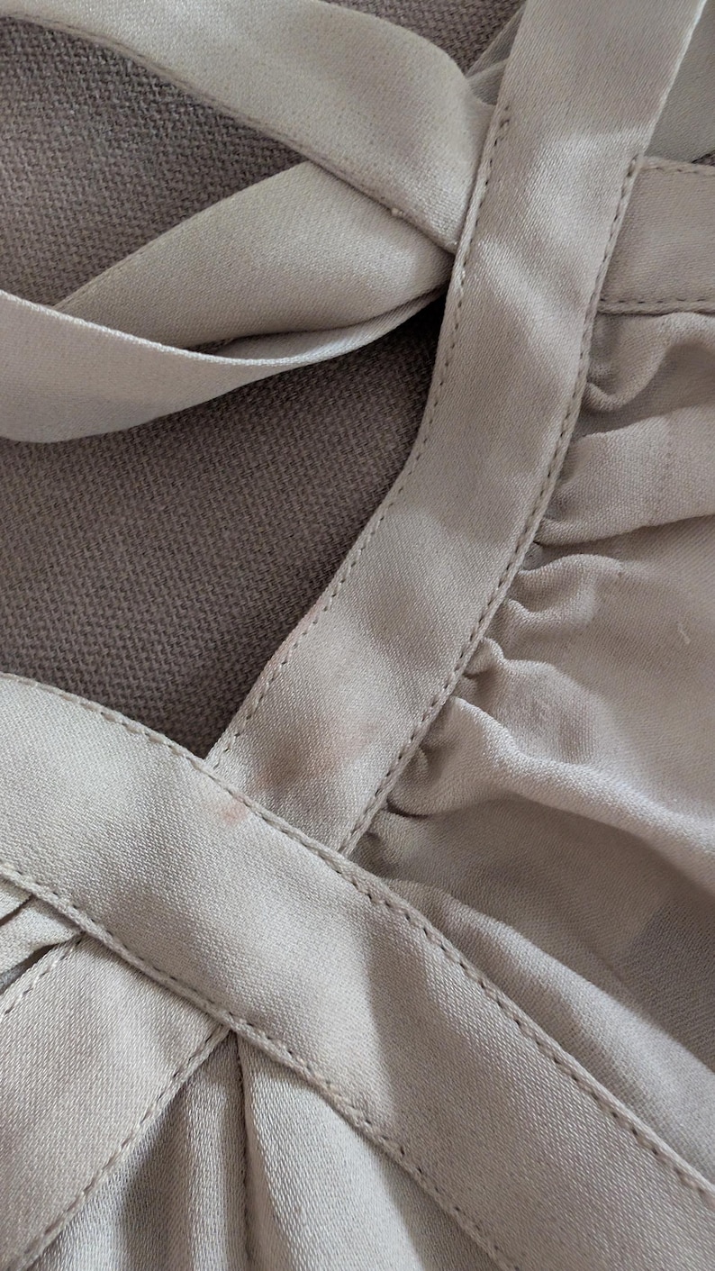 May include: A light beige garment featuring a rounded neckline with thick, stitched fabric bands that cross over each other. The fabric is gathered and pleated, creating soft folds and a textured appearance across the surface of the piece.
