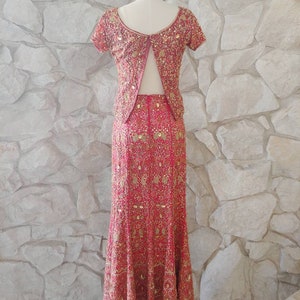 May include: A two-piece formal outfit in a vibrant red hue, adorned with intricate gold embroidery. The ensemble includes a short-sleeved top and a floor-length skirt, both embellished with detailed patterns. The outfit is displayed on a mannequin against a textured stone wall.
