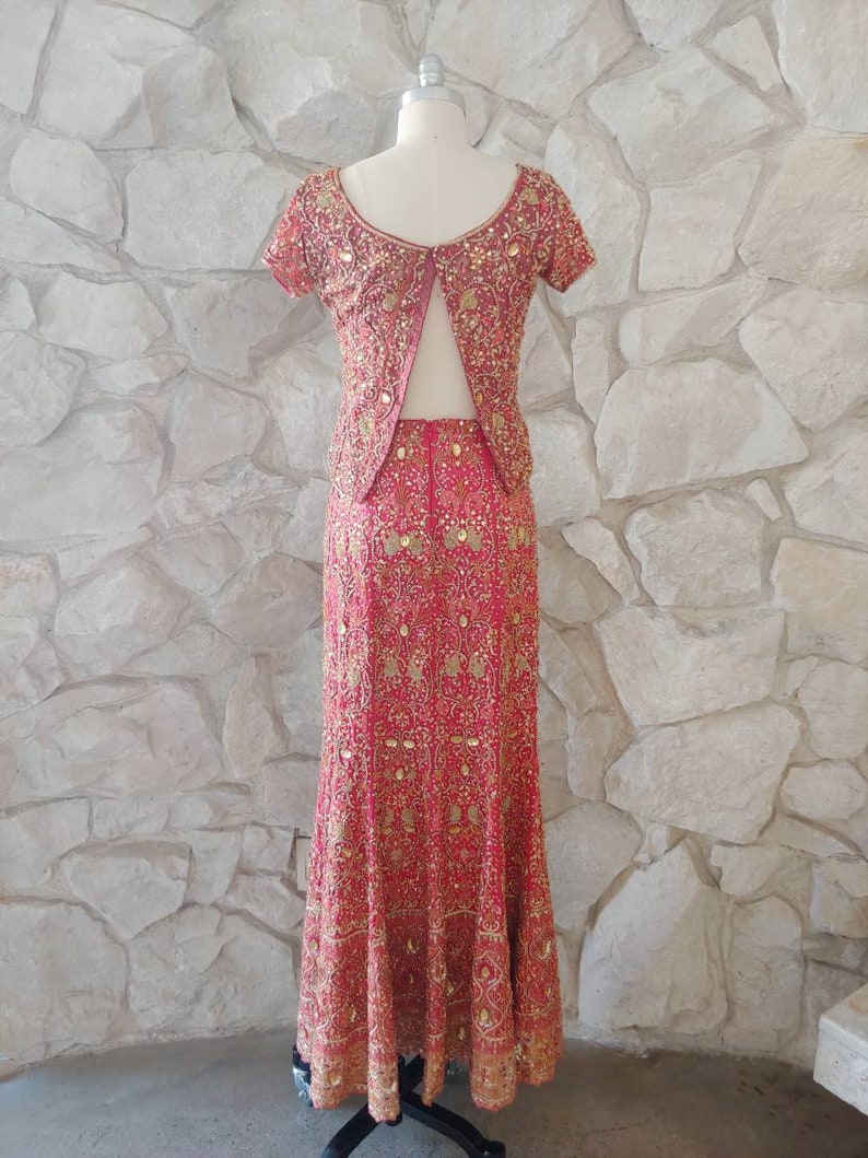 May include: A two-piece formal outfit in a vibrant red hue, adorned with intricate gold embroidery. The ensemble includes a short-sleeved top and a floor-length skirt, both embellished with detailed patterns. The outfit is displayed on a mannequin against a textured stone wall.