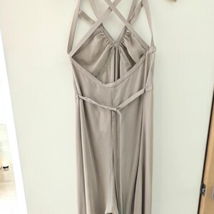 May include: A light beige jumpsuit hanging on a wooden hanger. The garment features a halter neckline with thin straps that cross over the open back, a gathered bodice, and a thin tie belt at the waist. The fabric has a smooth, satin-like finish.