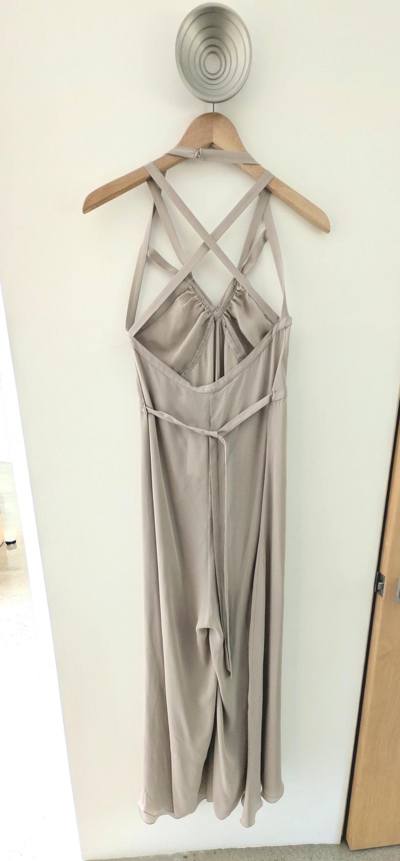 May include: A light beige jumpsuit hanging on a wooden hanger. The garment features a halter neckline with thin straps that cross over the open back, a gathered bodice, and a thin tie belt at the waist. The fabric has a smooth, satin-like finish.