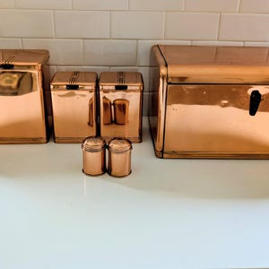 Pode incluir: Um conjunto de recipientes de cozinha em cobre polido, incluindo uma caixa de pão retangular grande, dois potes altos, dois potes quadrados menores e dois saleiros cilíndricos. Cada peça apresenta um acabamento metálico e design simples.