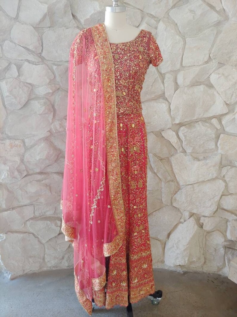 May include: A red and gold embroidered formal dress with a matching pink sheer scarf. The dress features intricate gold detailing and a short-sleeved design. The scarf is draped over the shoulder, adding a touch of elegance.