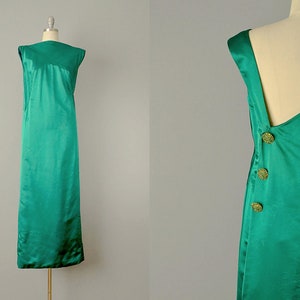 May include: Emerald green sleeveless dress with a round neckline and a long, straight silhouette. The back features a V-neck design with three decorative gold buttons. The dress is made of a shiny, satin-like fabric.