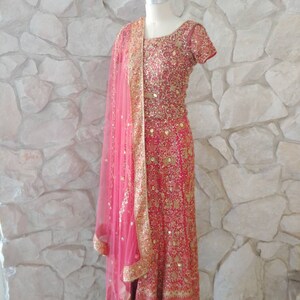 May include: A vibrant pink Indian dress with intricate gold embroidery and embellishments. The dress features a matching pink sheer scarf with gold trim. The dress is displayed on a mannequin against a textured stone wall.