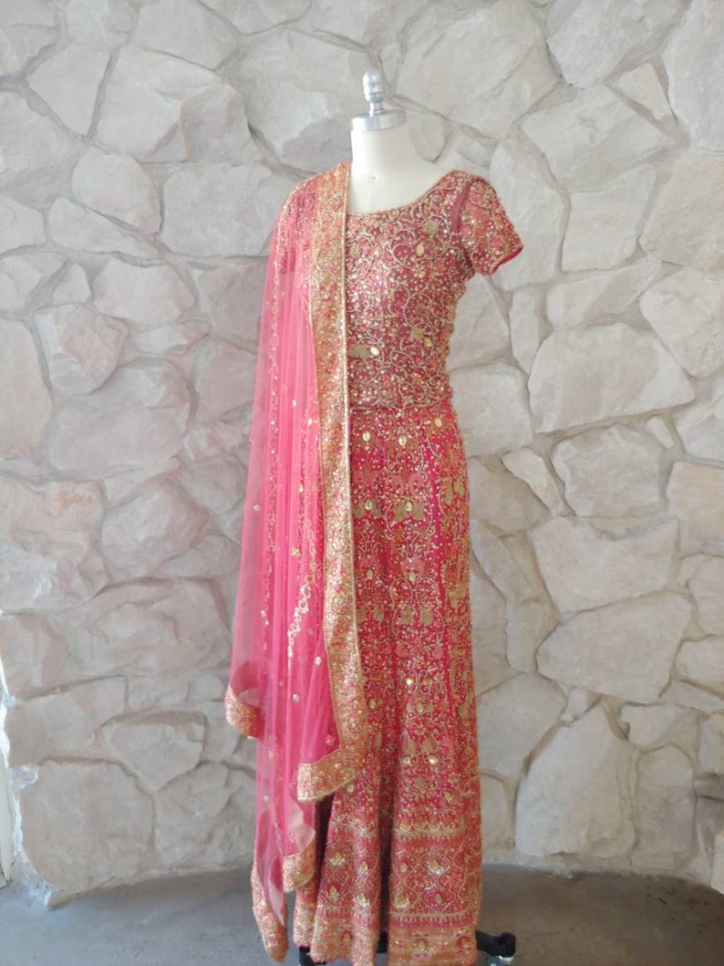 May include: A vibrant pink Indian dress with intricate gold embroidery and embellishments. The dress features a matching pink sheer scarf with gold trim. The dress is displayed on a mannequin against a textured stone wall.