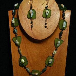 Puede incluir: Un collar y pendientes de vidrio verde con conchas marinas. El collar está hecho de cuentas de vidrio verde con conchas marinas incrustadas en cada cuenta. Los pendientes están hechos de vidrio verde con una concha marina incrustada en cada pendiente.