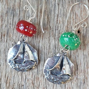 May include: A pair of silver earrings featuring a sailboat design. Each earring has a colorful glass bead with white dots. One bead is red, the other is green. The earrings hang from silver hooks.