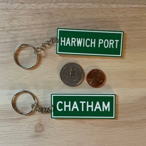 May include: Two green and white keychains with the words "Harwich Port" and "Chatham" on them. The keychains are on a wooden surface with two coins.