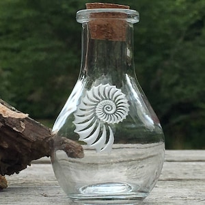 May include: A clear glass bottle with a cork stopper and a white etched spiral design. The bottle is sitting on a wooden surface.