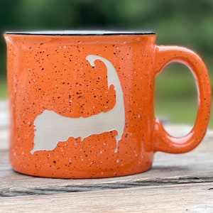 May include: Orange ceramic mug with a white outline of Cape Cod. The mug has a speckled orange glaze.