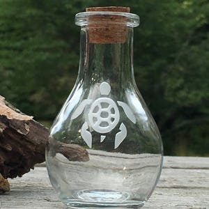 May include: A clear glass bottle with a cork stopper and a white etched sea turtle design. The bottle is sitting on a wooden surface.