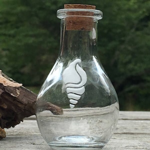 May include: A clear glass bottle with a cork stopper and a white seashell design. The bottle is empty and sits on a wooden surface, with a blurred green background.