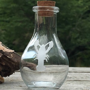 May include: A clear glass bottle with a cork stopper and a white etched design of a lobster. The bottle is sitting on a wooden surface.