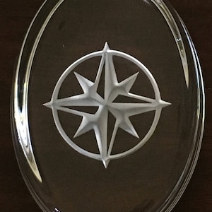 May include: Clear, oval-shaped glass paperweight with a frosted white compass rose design. The compass rose features eight points within a circular frame. The paperweight is approximately 4 inches long and 3 inches wide.