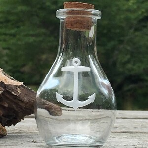 May include: A clear glass bottle with a cork stopper and a white anchor design. The bottle is empty and sits on a wooden surface, with a blurred green background.
