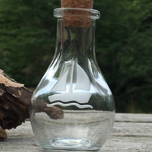 May include: A clear glass bottle with a cork stopper. The bottle features a white sailboat design etched on the front. The bottle is sitting on a wooden surface with a blurred green background.