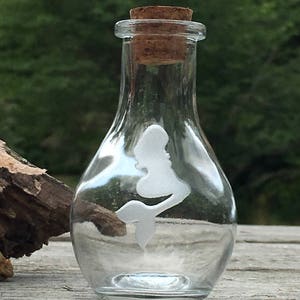 May include: A clear glass bottle with a cork stopper and a white silhouette of a mermaid etched on the side.