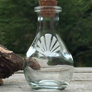 May include: A clear glass bottle with a cork stopper and a white seashell design. The bottle is sitting on a wooden surface.