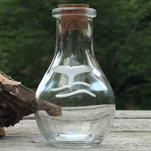 May include: A clear glass bottle with a cork stopper. The bottle features a white whale tail design above two wavy lines. The bottle is sitting on a wooden surface with a blurred green background.
