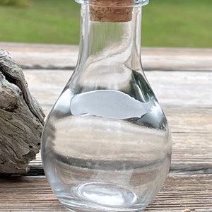 May include: A clear glass bottle with a cork stopper. The bottle is round and has a narrow neck. It is sitting on a wooden surface.