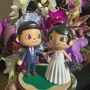 May include: A wedding cake topper featuring two cartoon figures, a bride and groom, holding hands. The groom wears a blue suit and the bride a white wedding dress. The base is round with green and brown accents, set against a backdrop of colorful flowers.