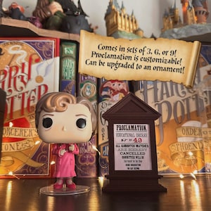 Może przedstawiać: Różowa i brązowa figurka Funko Pop! przedstawiająca kobietę z brązowymi włosami i okularami. Figurka stoi na przezroczystej plastikowej podstawie. Figurka stoi przed półką z książkami o Harrym Potterze i tabliczką z napisem "PROKLAMACJA. DEKRET EDUKACYJNY Nr 49 WSZYSTKIE MECZE QUAFFLE SĄ NINIEJSZYM ANULOWANE MIOTŁY ZOSTANĄ przekazane WYSOKIEJ INKWIZYTORCE DO BEZPIECZNEGO PRZECHOWANIA."
