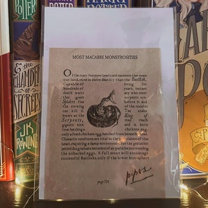 Puede incluir: Primer plano de una tarjeta marrón con el texto "Most Macabre Monstrosities" y una descripción del Basilisco, una criatura mítica del universo de Harry Potter. La tarjeta está sobre una mesa con libros de Harry Potter en el fondo.