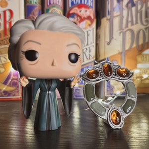 May include: A Pop Funko figure of a woman with gray hair wearing a dark green robe and holding a wand. The figure is standing in front of a bookshelf with Harry Potter books. A silver ring with three amber stones is in the foreground.