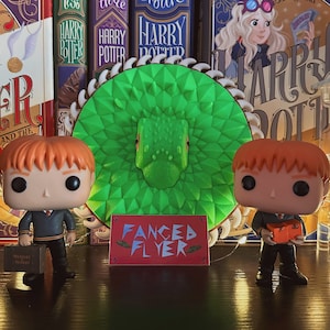 Puede incluir: Dos figuras Funko Pop de Harry Potter, una con una maleta marrón y la otra con un libro rojo, se encuentran frente a un juguete en forma de dragón verde y blanco. Las figuras están sobre una superficie de madera oscura con una tarjeta roja que dice "Fanced Flyer" delante de ellas. Las figuras están rodeadas de libros de Harry Potter.