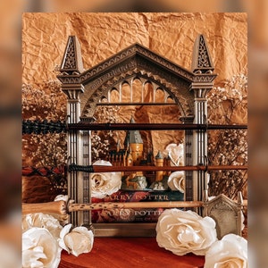 Pode incluir: Uma exibição decorativa com livros de Harry Potter, varinhas e um castelo de Hogwarts em miniatura. A exibição é montada sobre um fundo castanho com rosas brancas e flores secas. As varinhas são exibidas numa prateleira.