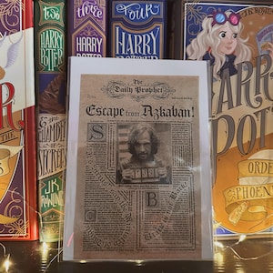 May include: A print of The Daily Prophet newspaper with the headline "Escape from Azkaban!" featuring a photo of Sirius Black. The print is displayed in front of several Harry Potter book covers, including "Harry Potter and the Chamber of Secrets."