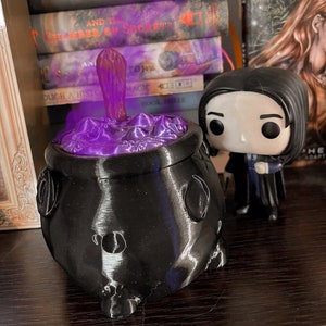 May include: A black cauldron with a purple glow emanating from the top. The cauldron is sitting on a wooden surface. A small figurine of a character from the Harry Potter series is standing next to the cauldron.