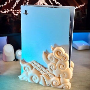 May include: A white PlayStation 5 console with a cloud-shaped stand. The stand is made of a light beige plastic and features intricate swirling designs.